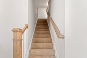 A carpeted staircase with wooden handrails on both sides, leading up to a landing area. Walls are painted white.