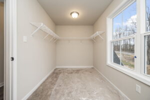 Empty walk-in closet with hanging wire shelves, beige walls, ceiling light, and large window overlooking outdoor trees. Floor partially covered with plastic sheet.