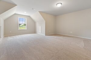 An empty, carpeted room with slanted ceilings, a single window letting in natural light, and a ceiling light fixture. The walls are painted light beige.