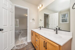 A bathroom with a large mirror, double sinks, wooden cabinets, and a door leading to an adjacent room with plastic covering the floor.