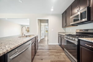 A modern kitchen with dark wood cabinets, granite countertops, stainless steel appliances, and a sink on the kitchen island. A hallway leads to other rooms.