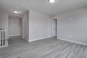A minimalistic empty room with light grey walls, a light fixture on the ceiling, and wood-patterned grey flooring. A white door and a closed-off staircase are visible.