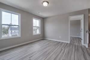 A vacant room with light gray wooden flooring, beige walls, two windows, and a ceiling light. An open door leads to another empty room.