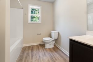 A bathroom with a bathtub and shower combination, a white toilet, a small window with a view of greenery, wood-style flooring, and a countertop with a black vanity.