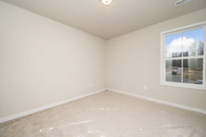 A vacant room with beige carpet, beige walls, and a white-trimmed window showing a view outside. The carpet is partially covered with clear plastic. Ceiling light is visible. Room is empty.