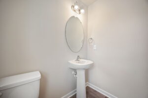 A small bathroom with a pedestal sink, oval mirror, light fixture above the mirror, toilet, and towel ring. The walls are painted light gray and the floor is made of wood.