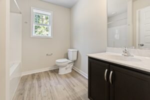 A clean, well-lit bathroom with a wooden floor, white toilet, marble countertop, dark wood vanity, and a tiled bathtub with a window above it.