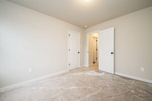 A room with beige walls, a beige carpet partly covered in plastic, and a small closet with double doors. The room connects to a hallway through an open door with a white frame.