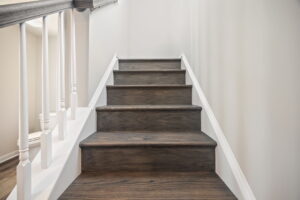 A set of wooden stairs with a white handrail on the left side, leading upwards in an indoor setting with neutral-colored walls.