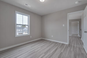 An empty room with light gray walls, a window with an outside view, light wood flooring, and a door leading to another room.