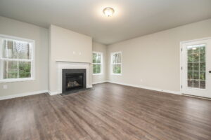 An empty living room with light-colored walls, wood flooring, a fireplace, two windows, a glass door, and overhead lighting.