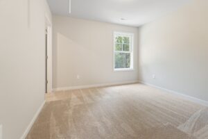 A small, empty room with beige walls and carpet. There is a single window with a view of trees and a door on the left side.