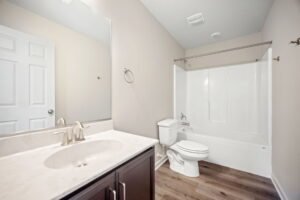 A bathroom features a sink with a mirror above, a toilet, and a bathtub with a shower curtain rod, all with light-colored walls and wooden flooring.