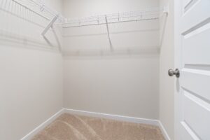 An empty walk-in closet with white wire shelving on beige walls and beige carpet flooring. The door is white with a silver knob.