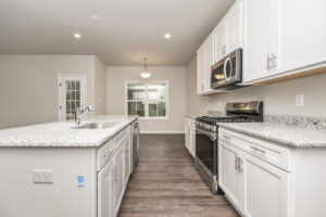 A modern kitchen with white cabinets, gray countertops, stainless steel appliances, and a central island sink. The room has hardwood flooring, recessed lighting, and a large window in the background.