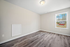 A small, empty room with light gray walls, a single overhead light, a window overlooking a construction site, and hardwood flooring. There is a square panel on one wall.