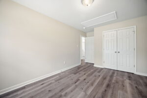 An empty room with beige walls, gray wood flooring, two white closet doors, and a white ceiling light. An open doorway leads to another space.