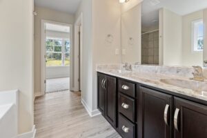 A bathroom featuring a double vanity with marble countertops, a large mirror, and dark wood cabinets. A doorway opens to a carpeted room with large windows, allowing natural light to enter.