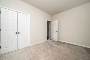 A small, empty room with beige walls and carpet, a double-door closet, and an open door leading to a hallway. Plastic sheets are spread on the floor.