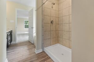 A bathroom with a tiled shower, wooden floor, and a doorway leading to another room with carpeting and a large window.