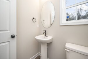 A small bathroom with an oval mirror above a white pedestal sink, a black faucet, minimal decor, and a window on the right side.