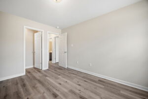 Empty room with light-colored walls and wooden flooring, showing two white doors leading to adjacent rooms. The ceiling light is on.