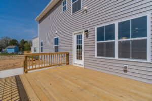 A wooden deck attached to the back of a beige house with multiple windows and a glass door, leading to a spacious backyard.