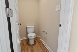 A small bathroom with a white toilet, beige walls, and wood flooring. The door is partially open, revealing a light switch on the wall and a toilet paper holder beside the toilet.