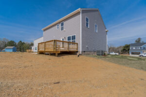 A two-story house with gray siding and a wooden deck in the backyard, situated on a barren and dry dirt lot with neighboring houses visible in the background.