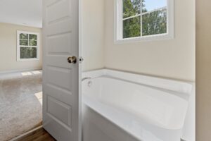A clean bathroom with a bathtub next to a partially open door, revealing a carpeted room with sunlight streaming through a window.