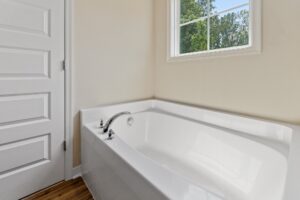 A white corner bathtub with a silver faucet is adjacent to a white-framed window with a view of green trees. A paneled white door is visible to the left.