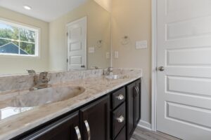 A bathroom features a double vanity with a marble countertop and dark wood cabinets, alongside a white door and a large wall mirror reflecting natural light from a window.