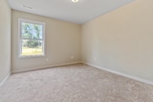 An empty room with beige carpet and light beige walls. A window lets in natural light, and a ceiling light fixture is visible.