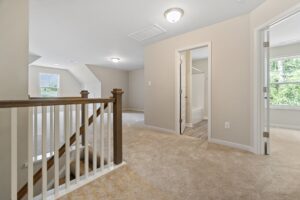 Carpeted upstairs hallway with wooden railing and light fixtures, leading to multiple rooms with white doors and a bathroom visible through one door. Large window provides natural light.