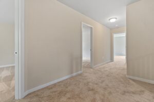 A hallway with beige walls and carpeted floor, leading to multiple doorways and lit by a ceiling light fixture.