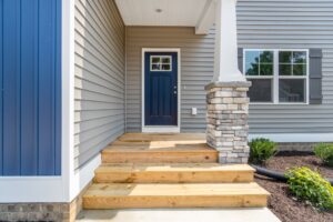 A front porch with wooden steps leading to a blue door, surrounded by light gray siding and a column with brick accents.
