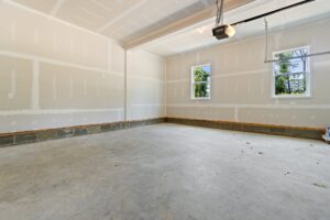 An empty, newly constructed garage with unpainted drywall, a concrete floor, two windows, and a ceiling-mounted garage door opener.