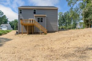 A two-story gray house with a wooden deck and stairs at the back, situated on a grassy lot with surrounding trees.