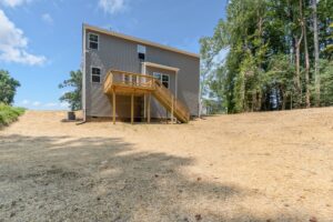 A two-story house with gray siding, a wooden deck, and stairs in the backyard, surrounded by a dirt and straw-covered ground with trees in the background.