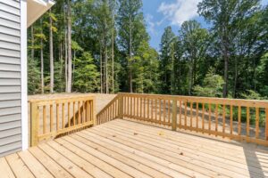 A wooden deck with railings overlooking a forested area behind a house. The sky is clear and blue, indicating a sunny day.