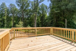 A wooden deck with railings overlooks a wooded area with tall trees under a clear sky.