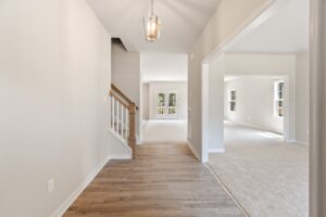 A bright, empty hallway with wooden flooring leads to a carpeted living area. A staircase with a wooden banister is on the left. A chandelier hangs from the ceiling. Doors and windows are visible ahead.