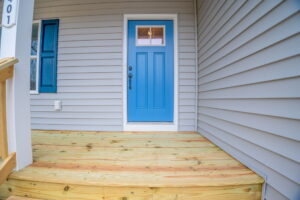 A wooden porch leads to a closed blue front door with a window panel, flanked by blue shutters on a grey house exterior.