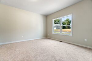 An empty room with beige walls, a large window, and carpeted flooring. Outside the window, there is a view of a grassy area and trees.