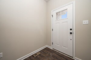 White entry door with a rectangular window at the top, set in a light-colored wall with a hardwood floor below. A light switch and floor vent are visible on the right and bottom respectively.