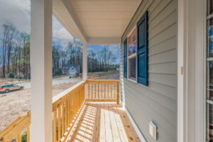 A wooden porch with railings attached to a light gray house with blue window shutters. The area is under construction with bare land and trees in the background. A car is parked on a dirt road.