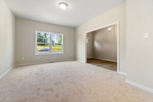 Empty room with beige carpet, light-colored walls, a ceiling light, and a window facing outside. An entryway connects to another room with wood flooring and a hanging light fixture.