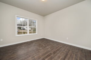 An empty room with light-colored walls, large window, and dark wood flooring. Outside the window is a view of trees and a house.