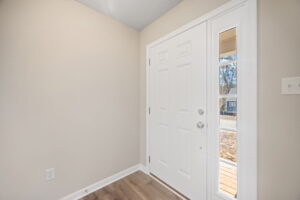 A white front door with an adjacent vertical window panel. The interior features beige walls and wood flooring. A light switch is visible on the right wall.