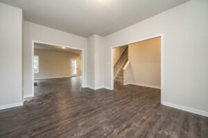 Empty room with dark wood flooring, beige walls, and open doorways. A staircase is visible in one doorway and another leads to an adjacent room with windows and more wood flooring.